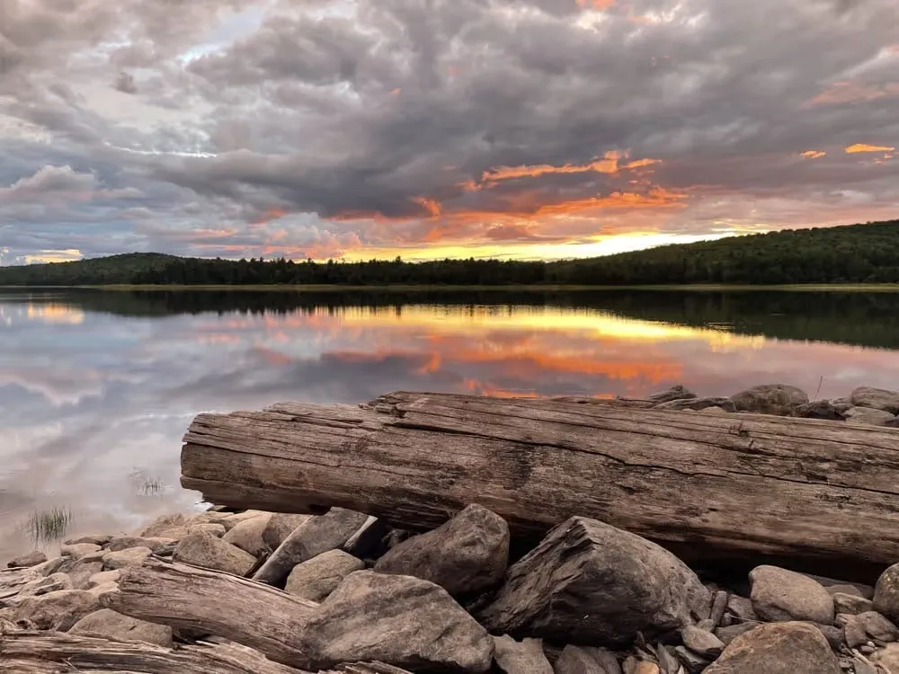 Driftwood log on a rocky shore beside a lake reflecting a sunset sky